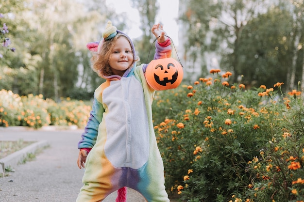 Śliczna mała dziewczynka w tęczowym kostiumie jednorożca na Halloween idzie po słodycze w dyniowym koszu