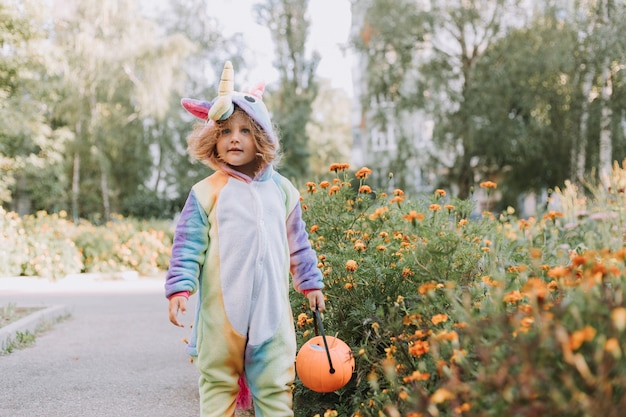 Śliczna mała dziewczynka w tęczowym kostiumie jednorożca na Halloween idzie po słodycze w dyniowym koszu