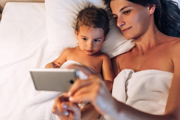 Śliczna mała dziewczynka z matką razem korzysta z telefonu w domu siedzi na łóżku Młoda kobieta i jej córeczka bawią się w sypialni Mama i córka używają gadżetu do nauki online