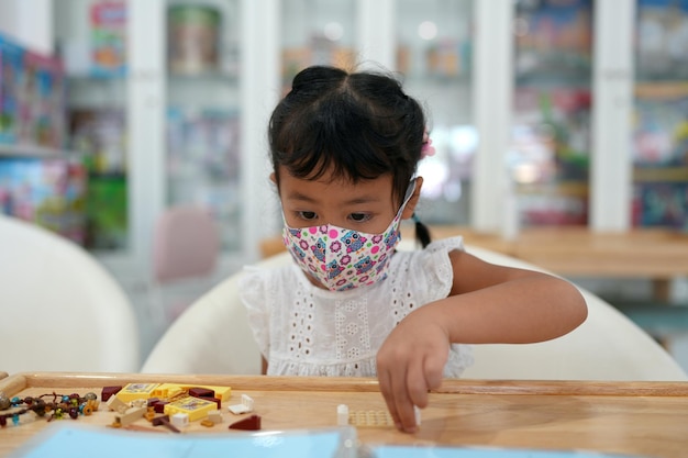 Śliczna mała dziewczynka z ochronną maską na twarz grająca w plastikowe klocki przy stole