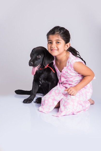Śliczna mała indyjska lub azjatycka dziewczyna bawi się szczeniakiem czarnego labrador retrievera, leżąc lub siedząc na białym tle