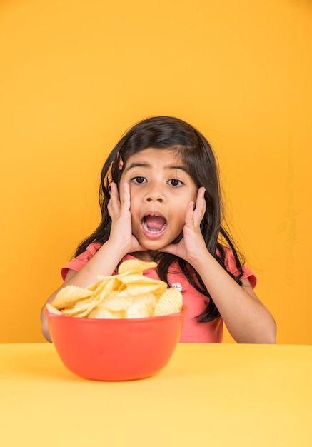 Śliczna mała indyjska lub azjatycka dziewczyna jedząca chipsy lub wafle ziemniaczane w dużej czerwonej misce, na żółtym tle
