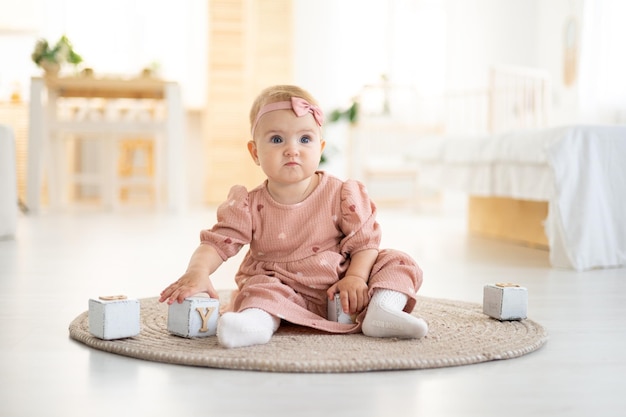 Śliczna mała zdrowa dziewczynka do roku życia w różowej sukience z naturalnej tkaniny siedzi na dywanie w salonie domu bawiąc się drewnianymi kostkami rozwój dziecka w domu
