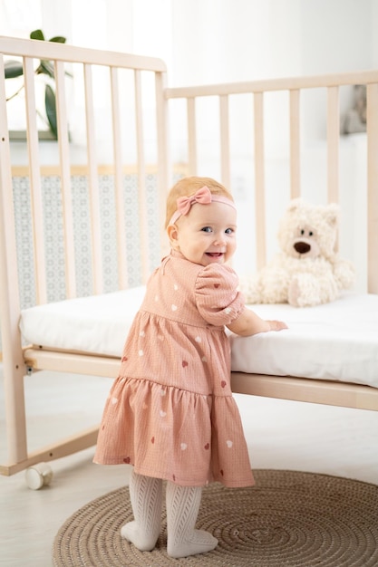 Śliczna mała zdrowa dziewczynka do roku życia w różowej sukience z naturalnej tkaniny stoi obok łóżeczka z misiami na białej pościeli w sypialni patrząc w kamerę