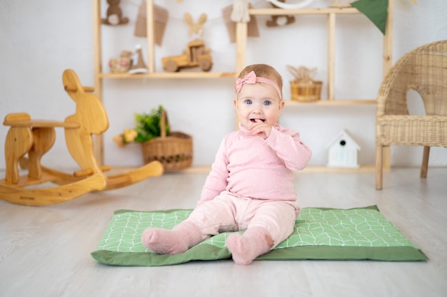 Śliczna mała zdrowa dziewczynka do roku życia w różowym garniturze z naturalnej tkaniny siedzi na dywaniku w pokoju dziecięcym z drewnianymi zabawkami edukacyjnymi patrząc w kamerę uśmiechając się