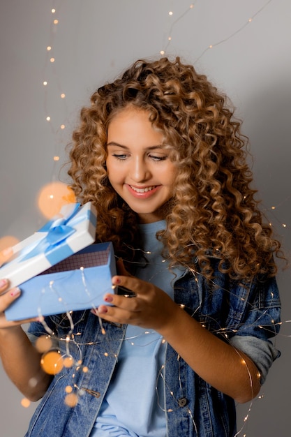 Śliczna młoda kobieta o niebieskich oczach i blond kręconych włosach w dżinsowych ubraniach trzyma w rękach pudełko na prezent