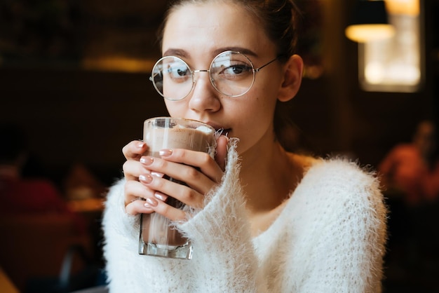 Śliczna młoda studentka w okularach pijąca pachnące latte w kawiarni odpoczywająca po studiach