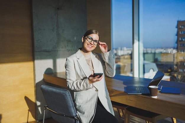 Śliczna pozytywna pracownica z długimi blond włosami w przypadkowych ubraniach patrząca na telefon podczas pracy nad nowym projektem biznesowym przy stole z laptopem i gadżetami w biurze
