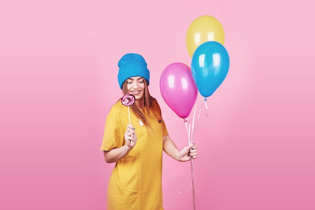 Śliczna śmieszna dziewczyna w błękitnej nakrętki portrecie trzyma lotniczych kolorowych balony i lizaka ono uśmiecha się na menchiach