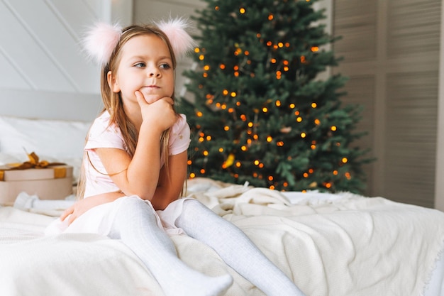 Śliczna smutna zabawna mała dziewczynka w różowej sukience z prezentem prezentowym siedzi na łóżku w pokoju z choinką Portret nieszczęśliwego dziecka w domku w okresie świąt Bożego Narodzenia Szczęśliwego Nowego Roku