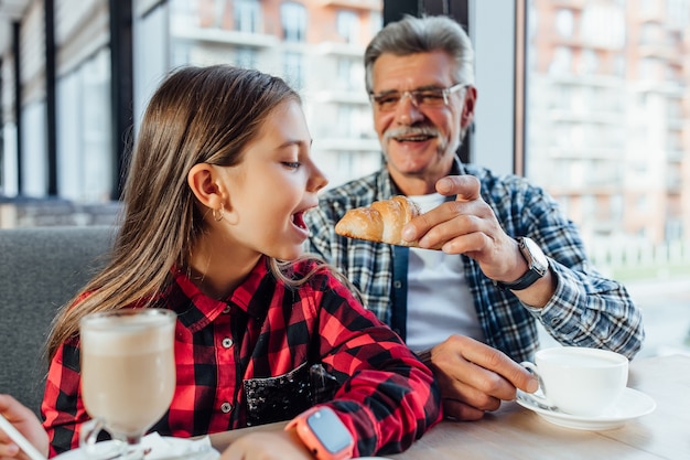 Śliczna Stylowa Wnuczka W Czerwonej Koszuli Próbująca Rogalika Jedzącego śniadanie Z Dziadkiem