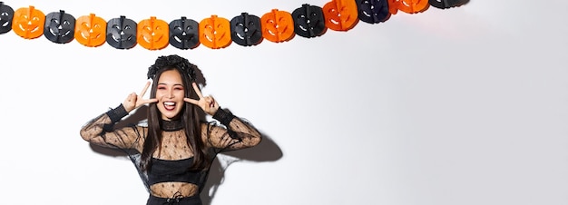 Śliczna szczęśliwa azjatycka kobieta ciesząca się imprezą z okazji halloween pokazującą gest pokoju i uśmiechnięta nosząca koszt czarownicy