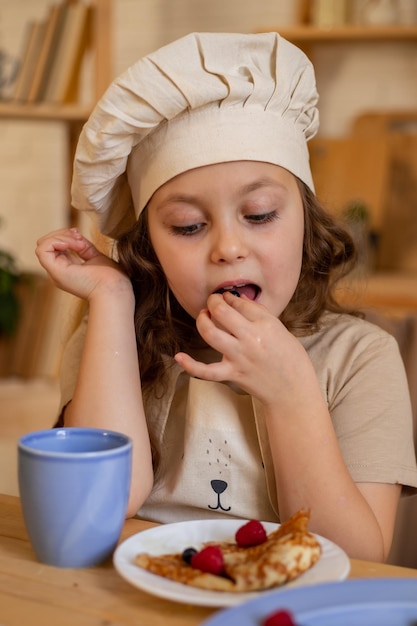 śliczna sześciolatka w kucharskiej czapce i fartuchu siedzi przy drewnianym stole jedząc naleśniki