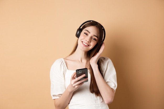 Śliczna uśmiechnięta dziewczyna słuchania muzyki w słuchawkach bezprzewodowych, za pomocą smartfona, stojąc na beżu.