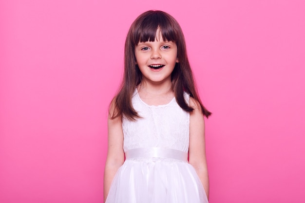 Śliczna uśmiechnięta dziewczyna ubrana w białą sukienkę, patrząc prosto z przodu z radosnym wyrazem twarzy, o ciemnych włosach, pozytywnym nastroju, odizolowana na różowej ścianie