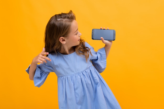 Śliczna uśmiechnięta dziewczyna w błękitnej sukni pokazuje telefon w horyzontalnej pozyci na kolorze żółtym z kopii przestrzenią