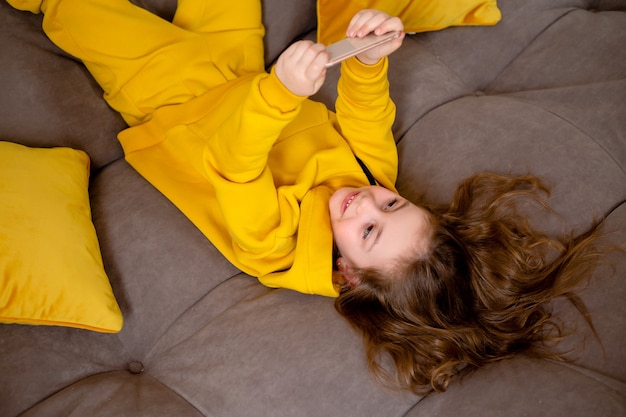 śliczna uśmiechnięta dziewczynka w żółtych ubraniach leży na plecach na łóżku i trzyma smartfon