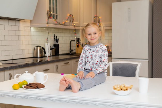 Śliczna uśmiechnięta mała blondynka w świątecznych ubraniach z nadrukiem siedzi w przytulnym kuchennym stylu życia