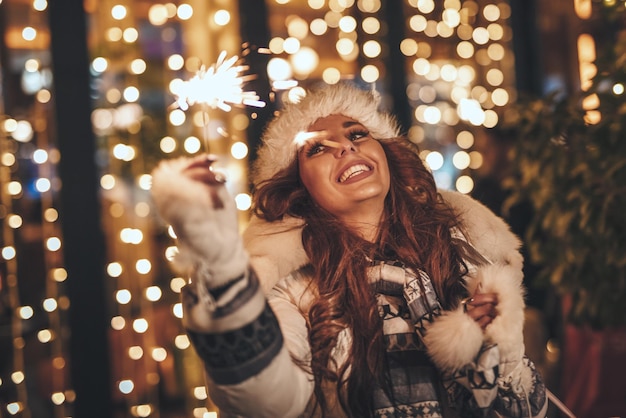 Śliczna wesoła dziewczyna z zimnymi ogniami bawi się w Wigilię Bożego Narodzenia na ulicy miasta z mnóstwem świątecznych świateł.