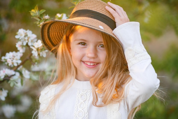 śliczna zabawna mała blondynka w słomkowym kapeluszu w pobliżu kwitnącego drzewa na wiosnę śmiejąca się