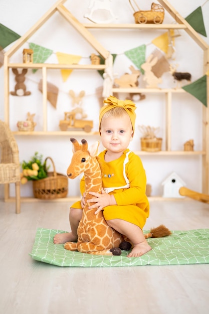 Śliczna zdrowa dziewczyna bawiąca się drewnianymi zabawkami edukacyjnymi w jasnym przytulnym pokoju dziecięcym w domu na tle drewnianego domu siedzącego na pluszowej żyrafie