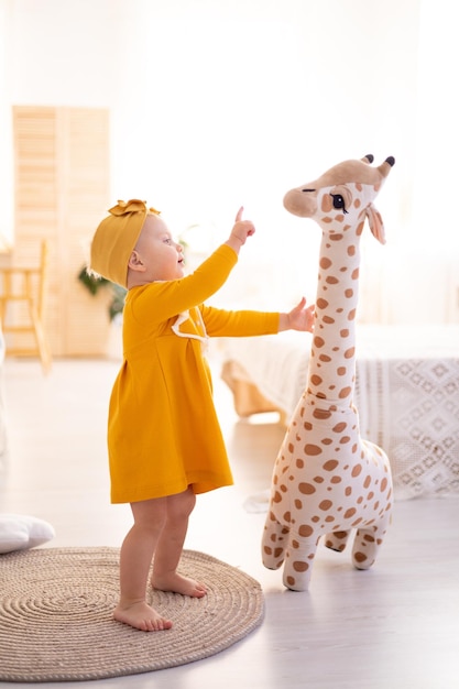 Śliczna zdrowa dziewczyna stoi na dywanie w jasnym salonie domu na tle łóżka bawiącego się wypchanymi tekstyliami domowymi żyrafy