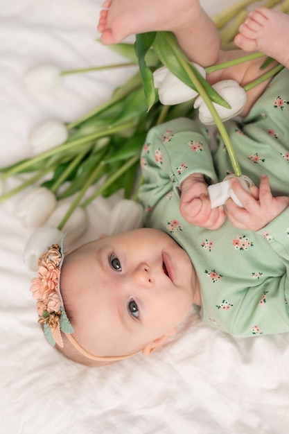 Śliczne Dziecko Z Kwiecistą Opaską I Zielonym Bawełnianym Body W Domu Na Białym łóżku Z Tulipanami. Spr