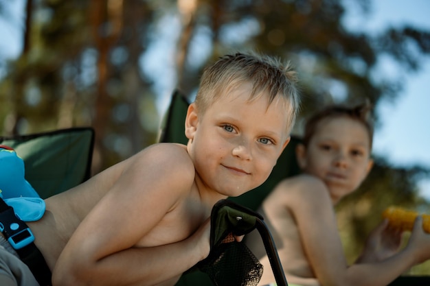 Śliczne kaukaskie chłopcy jedzą kukurydzę na plaży siedząc na krzesłach kempingowych, śmiejąc się, ciesząc się wakacjami