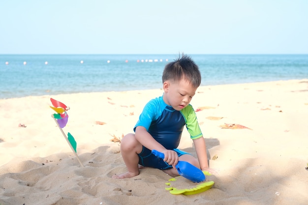 Śliczne Małe Azjatyckie Dziecko, Przedszkolak Siedzi I Bawi Się Zabawkami Na Plaży Tropikalnej