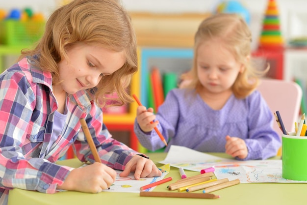 Śliczne małe dziewczynki siedzą przy stole i rysują ołówkami w swoim pokoju