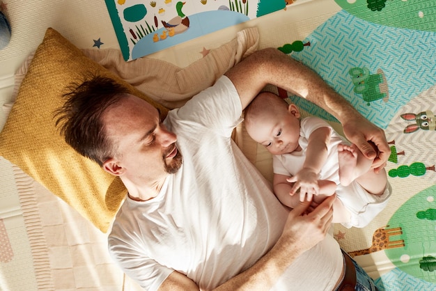 Śliczne radosne dziecko i jego ojciec w białej koszulce i dżinsach leżą na podłodze w jasnym pokoju i p