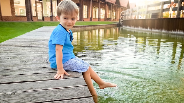 Śliczny 3-letni Chłopiec Malucha Siedzący Nad Brzegiem Rzeki W Małym Europejskim Miasteczku W Słoneczny Letni Dzień