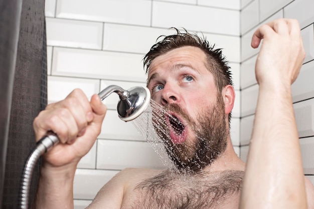 Śliczny brodaty mężczyzna śpiewa w łazience przy użyciu słuchawki prysznicowej z płynącą wodą zamiast mikrofonu