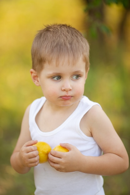 Śliczny Chłopak O Blond Włosach W Białej Koszulce Z Letnimi Cytrynami W Ogrodzie Pod Drzewem