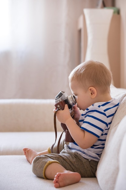 Śliczny chłopczyk siedzący na kanapie w salonie i bawiący się zabytkowym aparatem fotograficznym