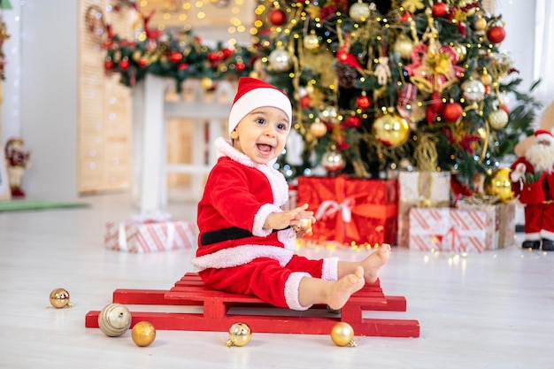 Śliczny chłopczyk w stroju Mikołaja siedzi na sankach pod świąteczną choinką z prezentami w salonie domu szczęśliwe dziecko świętuje Boże Narodzenie i Nowy Rok w domu