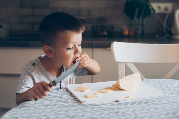 Śliczny chłopiec 4-5 lat z nożem tnącym ser na desce do krojenia na stole w kuchni