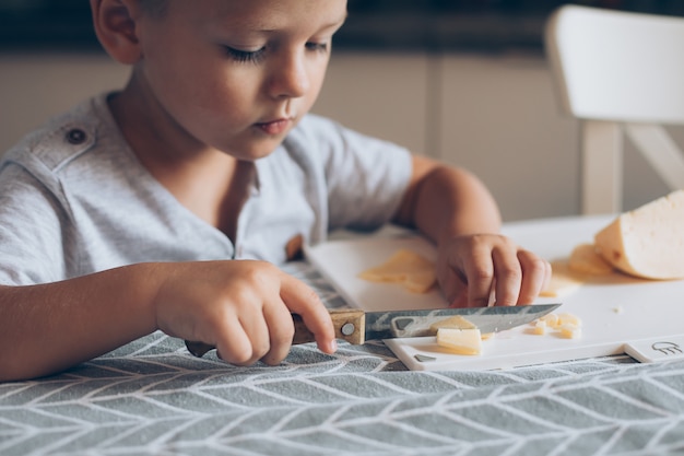 Śliczny chłopiec 4-5 lat z nożem tnącym ser na desce do krojenia na stole w kuchni