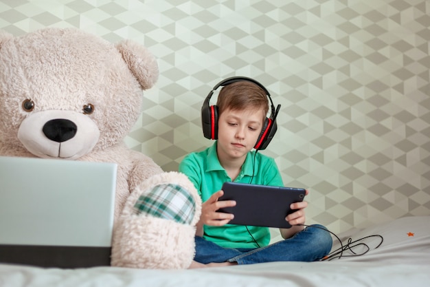 Śliczny dzieciak w hełmofonach bawić się na cyfrowej pastylce obok zabawkarskiego niedźwiedzia patrzeje w komputerze w domu.