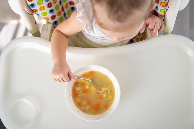 Śliczny Maluch Siedzący W Krzesełku I Jedzący Zupę Obiadową W Domowej Kuchni