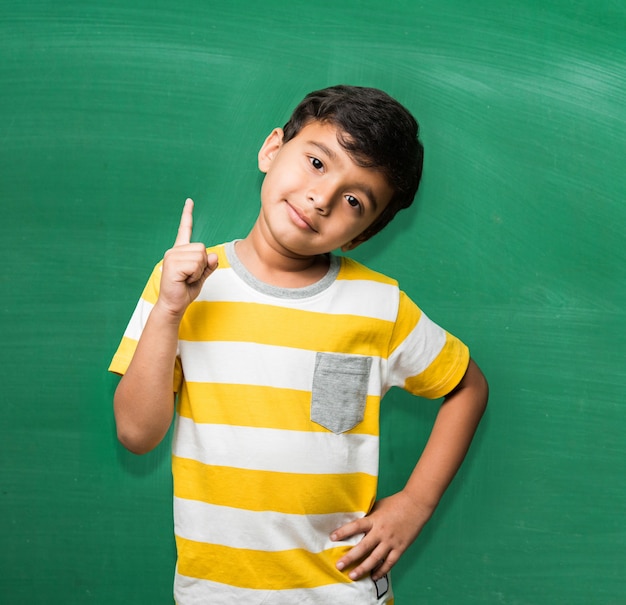 Śliczny mały chłopiec dziecko indyjskiej szkoły w ręku rozciągnięty poza na zielonej tablicy lub tle tablicy kredowej, trzymając książki, puchar zwycięstwa itp., na białym tle