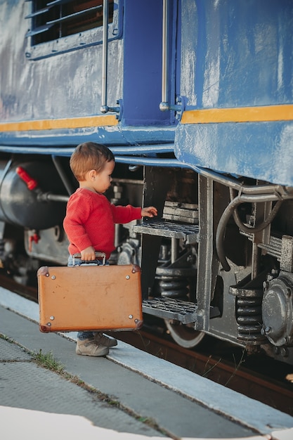 Śliczny mały chłopiec dziecko ubrany w czerwony sweter na stacji kolejowej w pobliżu pociągu z retro starą brązową walizką. Gotowy na wakacje. Młody podróżnik na peronie.