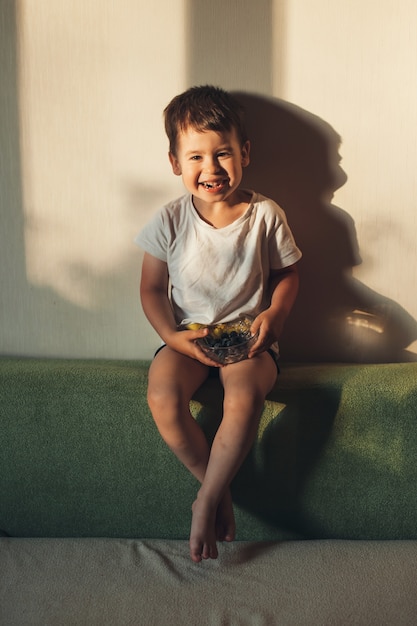 Śliczny mały chłopiec uśmiecha się do kamery podczas jedzenia jagód, siedząc na kanapie w domu