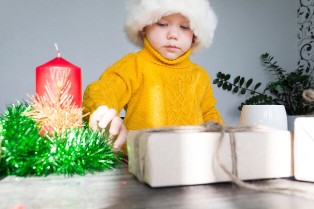Śliczny mały chłopiec w żółtym swetrze i czerwonym kapeluszu Świętego Mikołaja przy drewnianym stole z prezentami zapakowanymi w papier pakowy