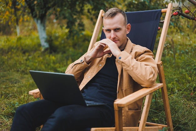 Śliczny mężczyzna w koszuli siedzi na krześle na zewnątrz w ogrodzie i pracuje na laptopie praca zdalna freelancer