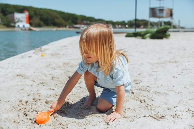Śliczny szczęśliwy blond chłopiec bawi się zabawkami plażowymi na piaszczystej plaży miejskiej
