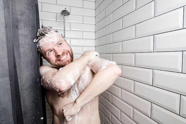 Śliczny szczęśliwy człowiek namydlony pianką myje się pod prysznicem pod bieżącą wodą