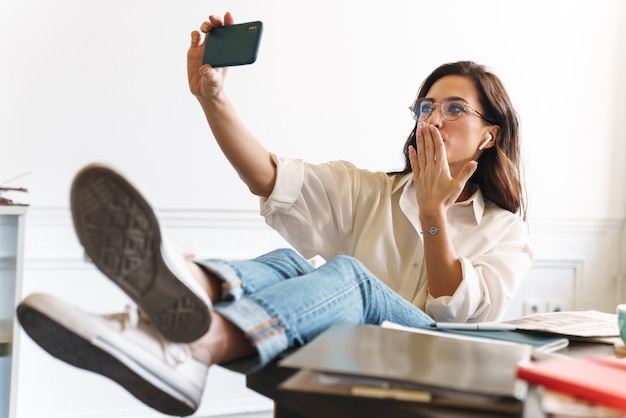 Śliczny Wesoły Młoda Brunetka Kobieta Relaks Przy Stole W Kawiarni W Pomieszczeniu, Biorąc Selfie