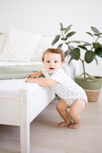 Śliczny zdrowy chłopczyk w przestronnej jasnej sypialni stoi obok łóżka z białą pościelą dziecko stoi z podparciem dziecko uczy się chodzić