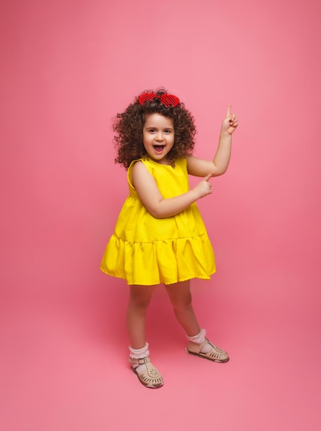 Słodka, atrakcyjna, radosna dziewczynka w żółtej sukience wskazująca palcem wskazującym na różowe pastelowe tło